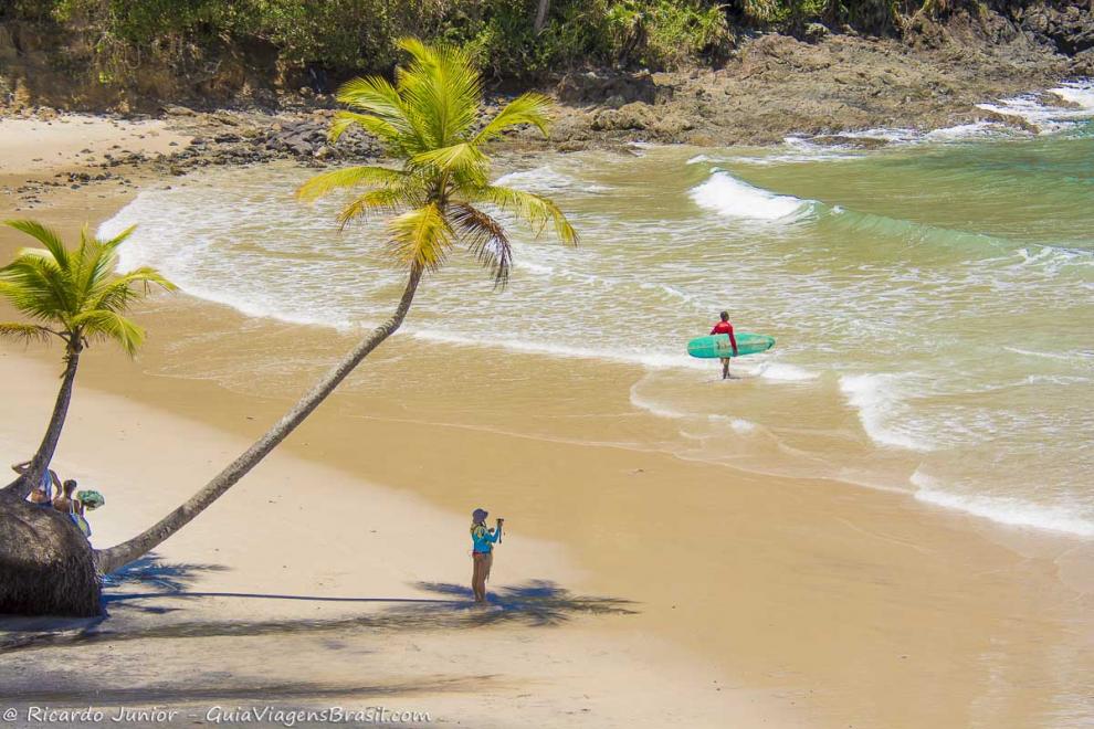 Imagem de um surfista chegando e menina na sombra de um coqueiro.